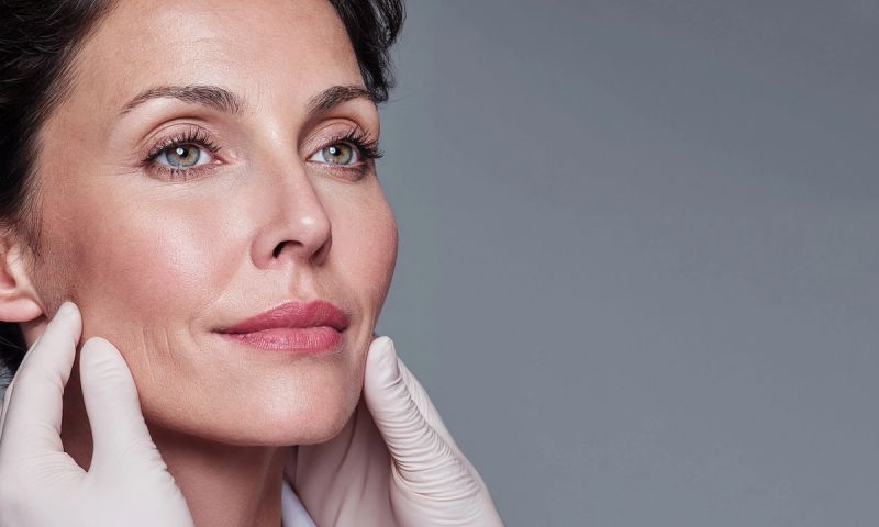 AI PROMPT (ALL): EDITORIAL BEAUTY SHOT OF A DERMATOLOGIST WEARING A WHITE COAT ASSESSING A WOMAN’S FACE (40S, GLOWING SKIN, SUBTLE FILLER ENHANCEMENTS IN CHEEKS AND LIPS)