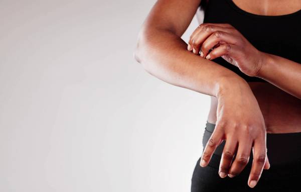 A woman in black activewear scratches her forearm against a white background.