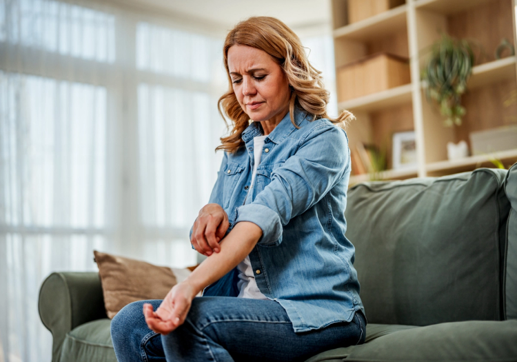 Woman sitting on a couch scratching her itchy forearm, wincing in discomfort