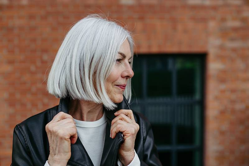 Portrait of stylish mature woman with gray hair on city street, outdoor. Older woman in leather jacket with soft smile on face.