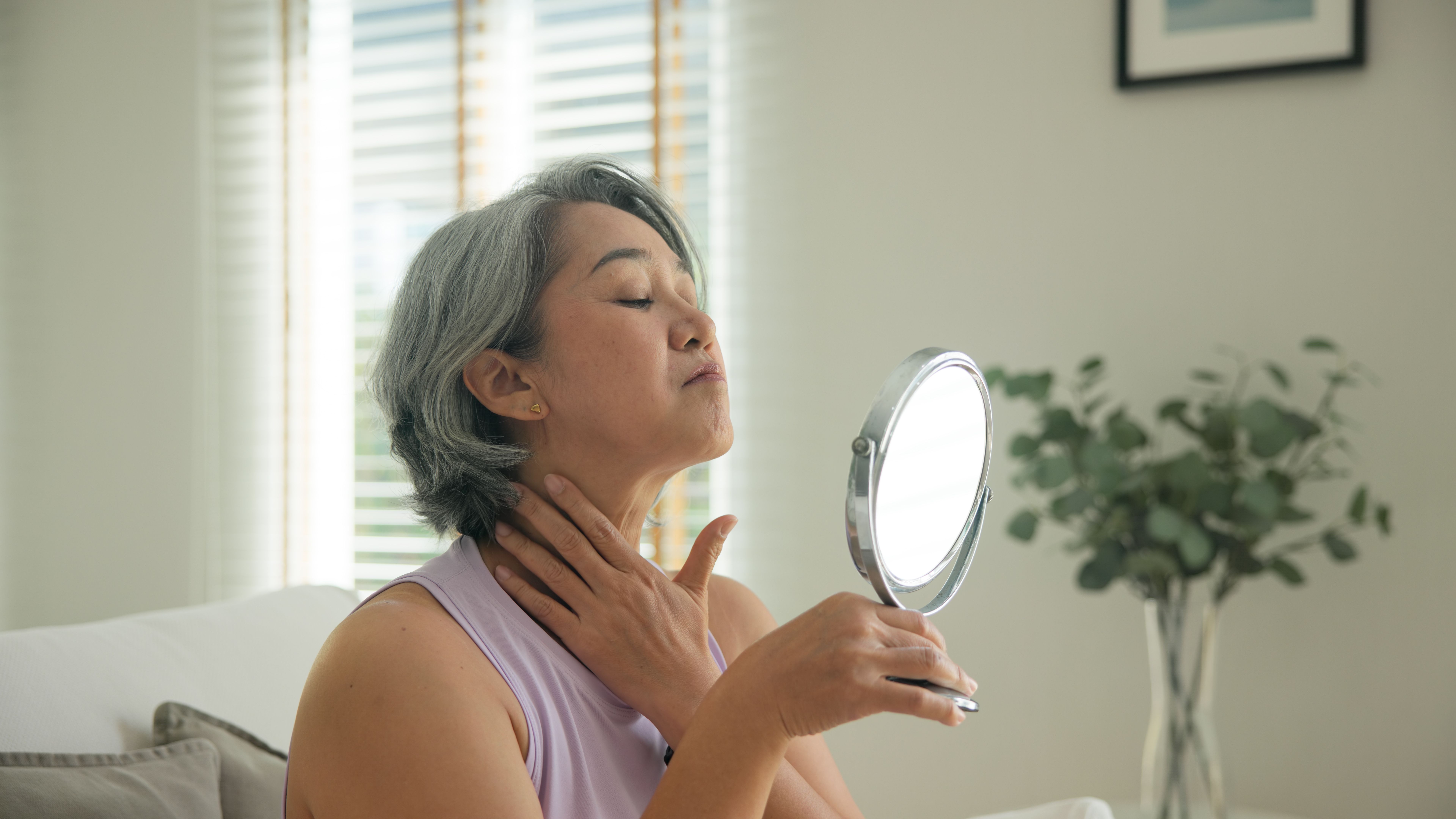 A woman with short gray hair holds a handheld mirror and touches her neck, chin tilted up.