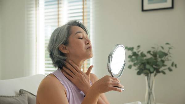 A woman with short gray hair holds a handheld mirror and touches her neck, chin tilted up.