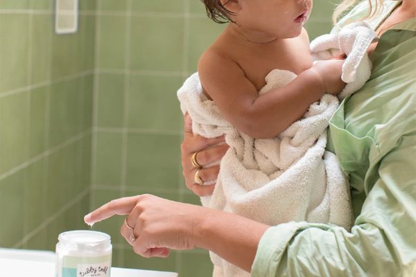 Parent holding a baby wrapped in a towel while reaching for a jar of baby skin-care cream on a bathroom counter.