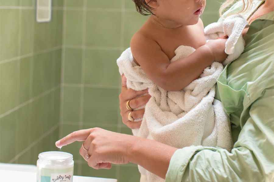 Parent holding a baby wrapped in a towel while reaching for a jar of baby skin-care cream on a bathroom counter.