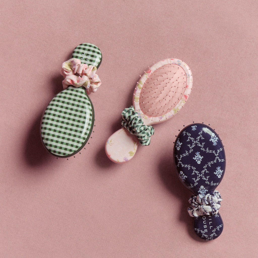 Three patterned hairbrushes from the Wet Brush x Hill House Home collaboration featuring gingham and floral prints, each styled with matching scrunchies on a pink background.