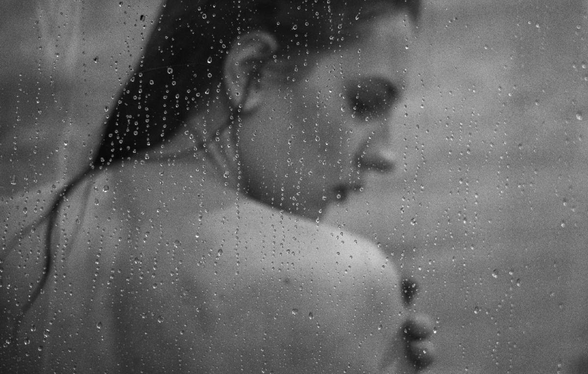 Black-and-white photo of a woman with wet hair seen through a water-droplet-covered shower door