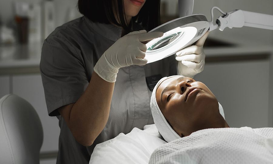 Beautician woman looking at client's face with lamp examining her skin. Relaxed young woman getting professional facial treatment in spa salon. Healing skin problems.