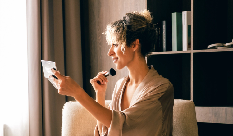 Woman sitting by a window applies blush with a makeup brush while looking into a compact mirror.