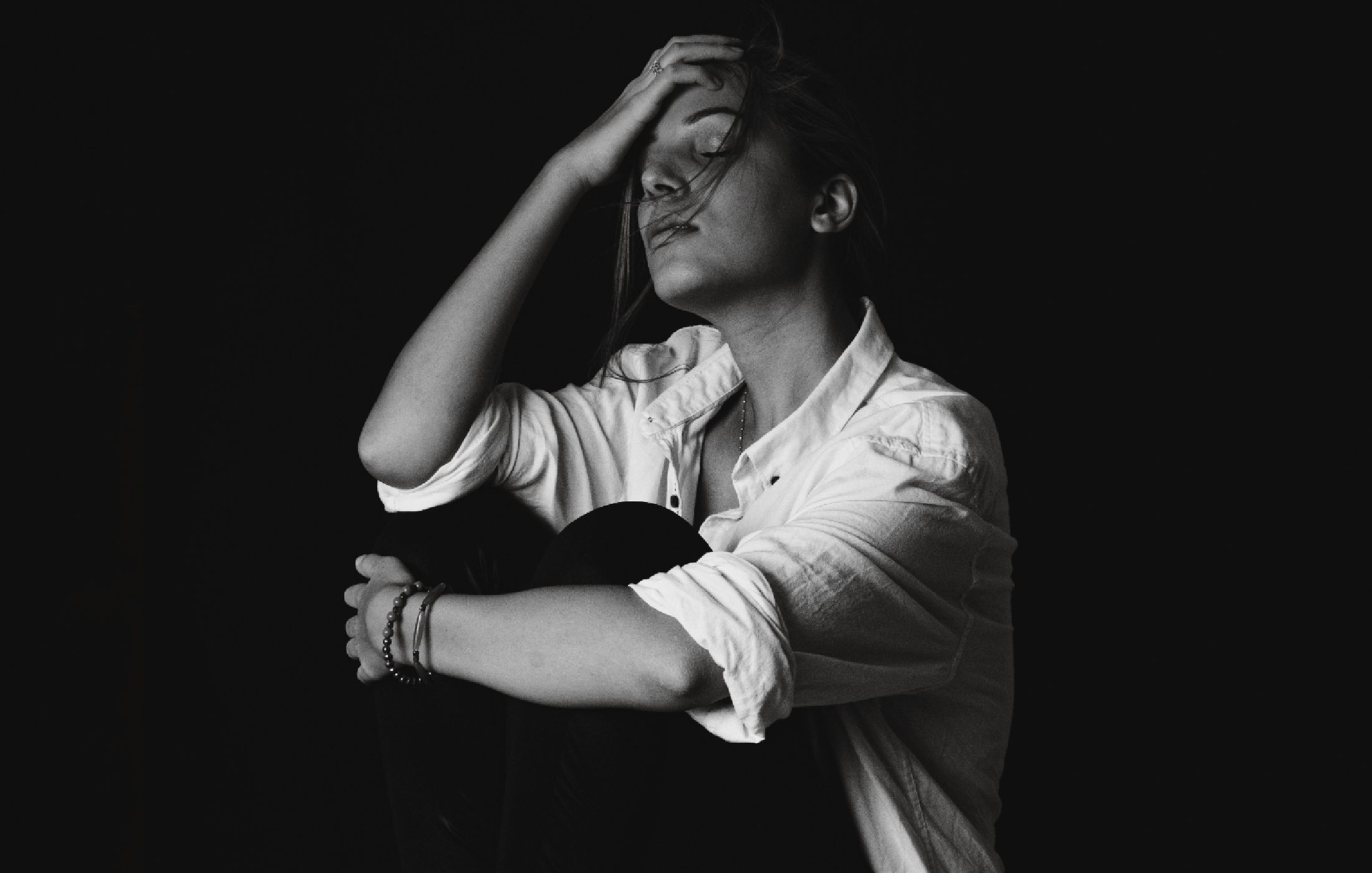 A woman sits with her head resting in her hand, photographed in black and white against a dark background