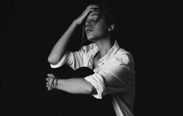 A woman sits with her head resting in her hand, photographed in black and white against a dark background