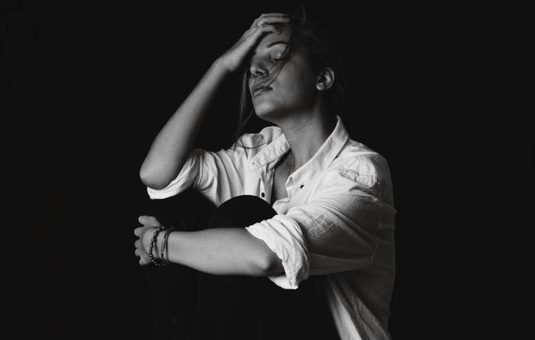 A woman sits with her head resting in her hand, photographed in black and white against a dark background