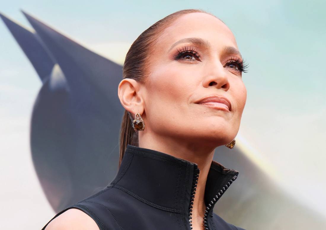 Jennifer Lopez wearing slicked-back hair, glowing makeup, statement earrings and a black high-neck top at an outdoor event.