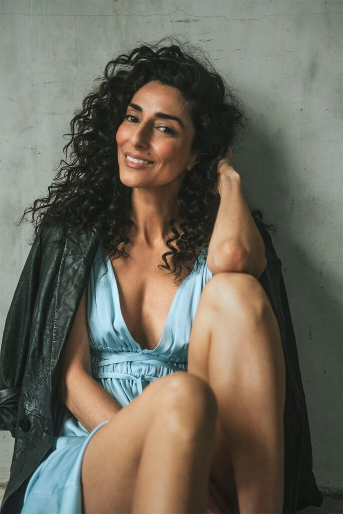 Necar Zadegan posing in a light blue dress with a leather jacket over her shoulders. 
