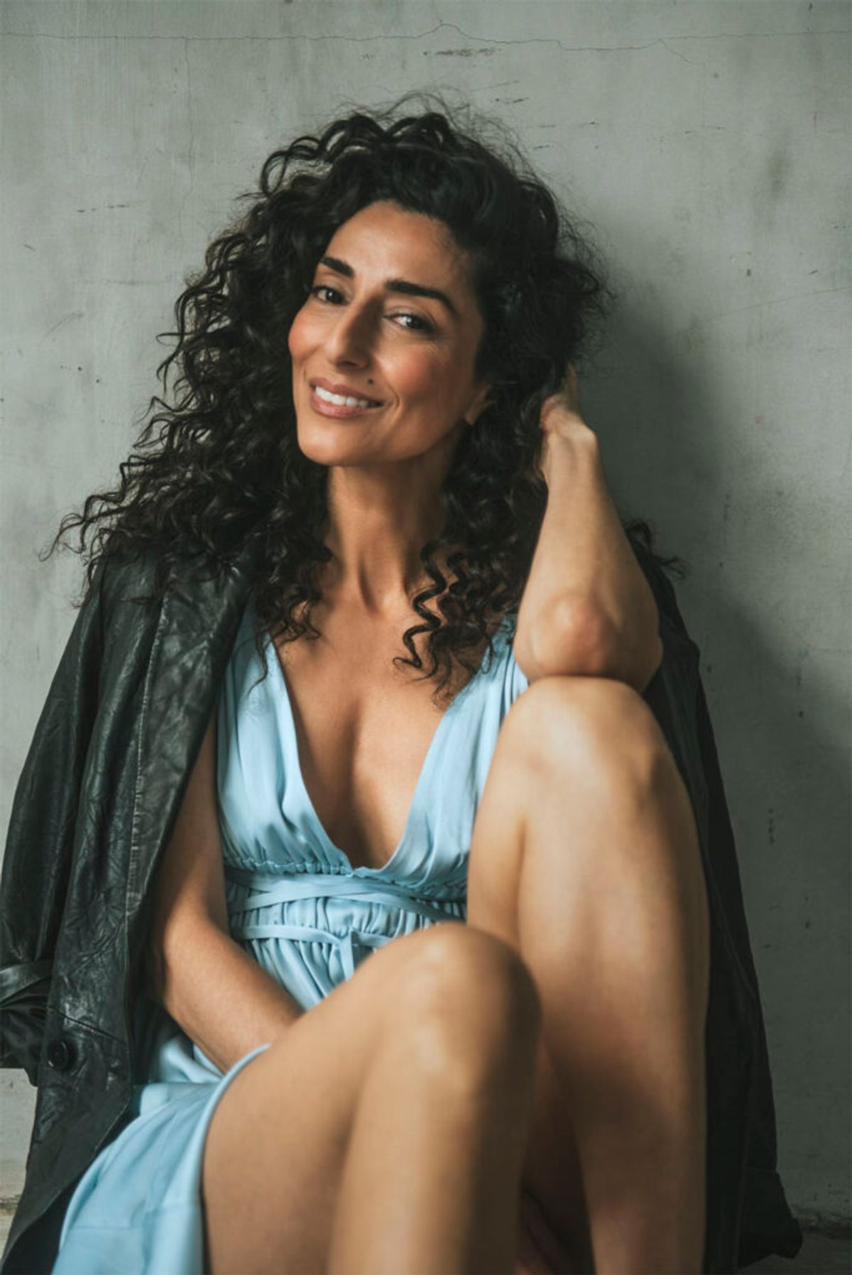 Necar Zadegan posing in a light blue dress with a leather jacket over her shoulders. 