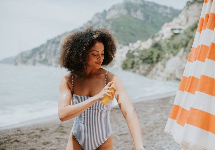 Woman applying spray sunscreen to her arm on a beach, with ocean and coastal cliffs in the background