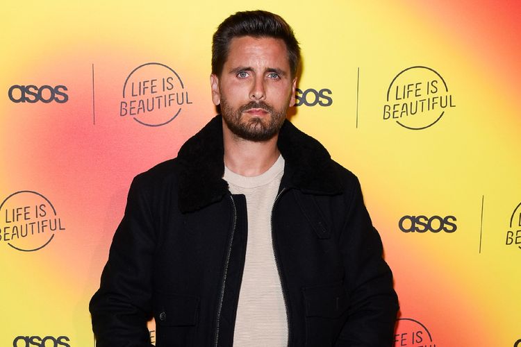 Scott Disick poses at a branded event in a black jacket against a yellow and orange backdrop featuring ASOS and Life Is Beautiful logos