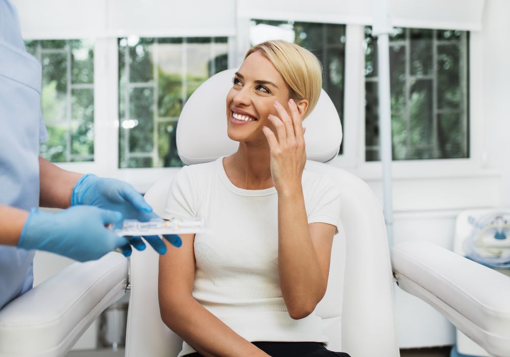 Woman smiling in a dermatology clinic chair as a provider prepares an injectable treatment, illustrating preventive aesthetics and subtle, natural-looking care.