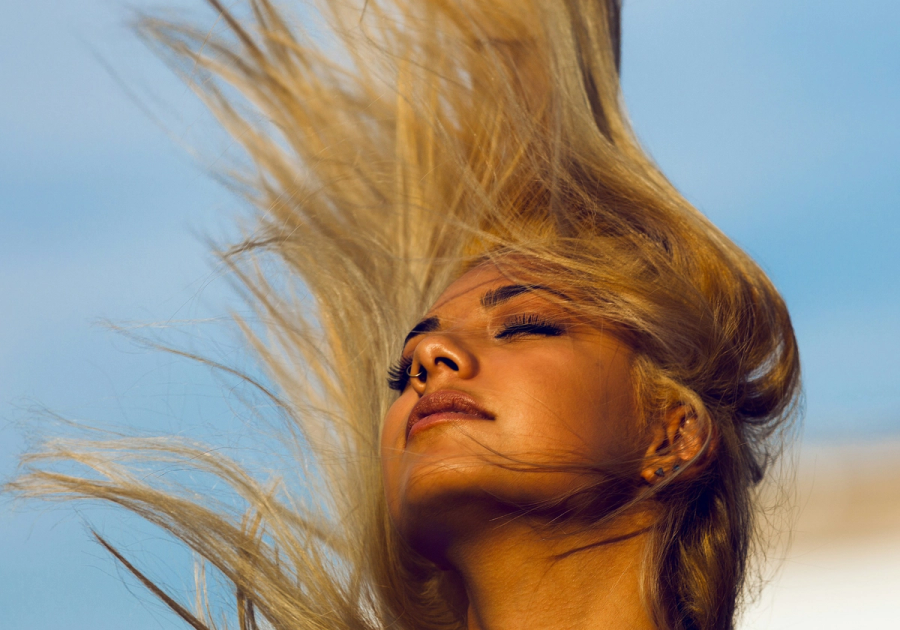 Close-up of a woman with eyes closed and head tilted back, her long wavy hair whipping upward against a clear blue sky.