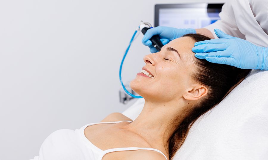 A middle-aged woman receives facial treatment in a professional beauty salon. A close-up cosmetologist manipulates a hydropeeling device to clean and rejuvenate facial skin.