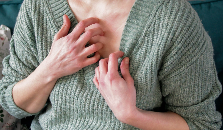 Close-up of a person in a green sweater scratching red, irritated skin on their chest.