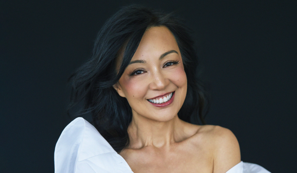 Ming-Na Wen smiles with her arms crossed, wearing an off-the-shoulder white dress against a dark backdrop.
