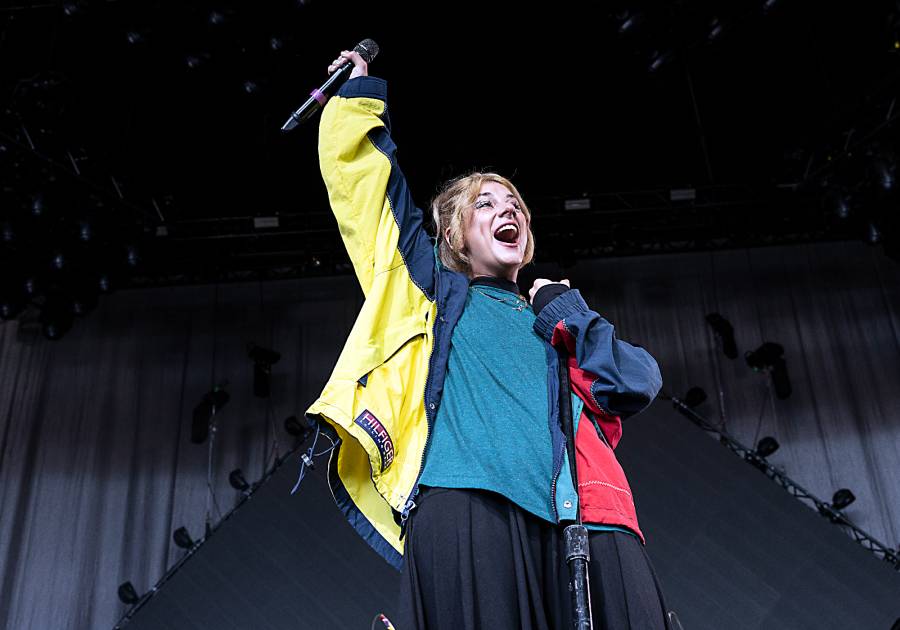 Singer-songwriter Jax smiles warmly at the camera, wearing a navy blue hoodie with her blonde hair pulled up in a top bun, against a neutral beige background.