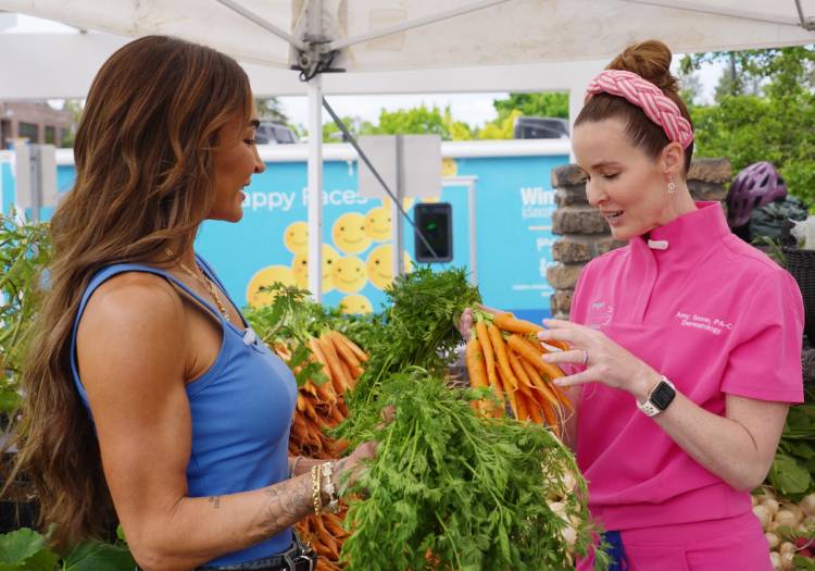 Food influencer Nicole Keshishian and dermatology PA-C Amy Snow examine fresh carrots at the Bend, Oregon farmers market in front of the Project Happy Face mobile dermatology unit