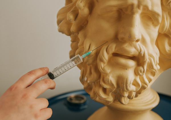 A hand holds a syringe with a needle inserted near the jawline of a golden bearded classical bust