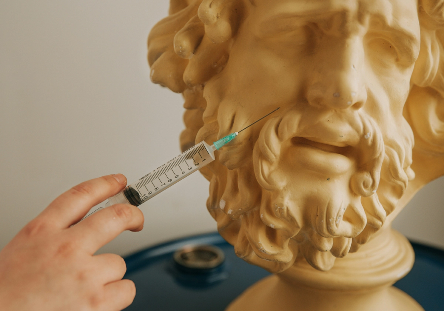 A hand holds a syringe with a needle inserted near the jawline of a golden bearded classical bust