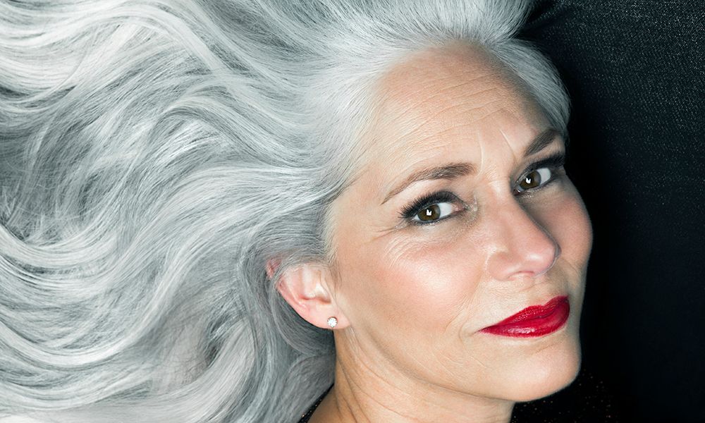 Woman in a glamorous portrait with her long, wavy, silver gray hair spread out behind her in front of a black background wearing red lipstick.