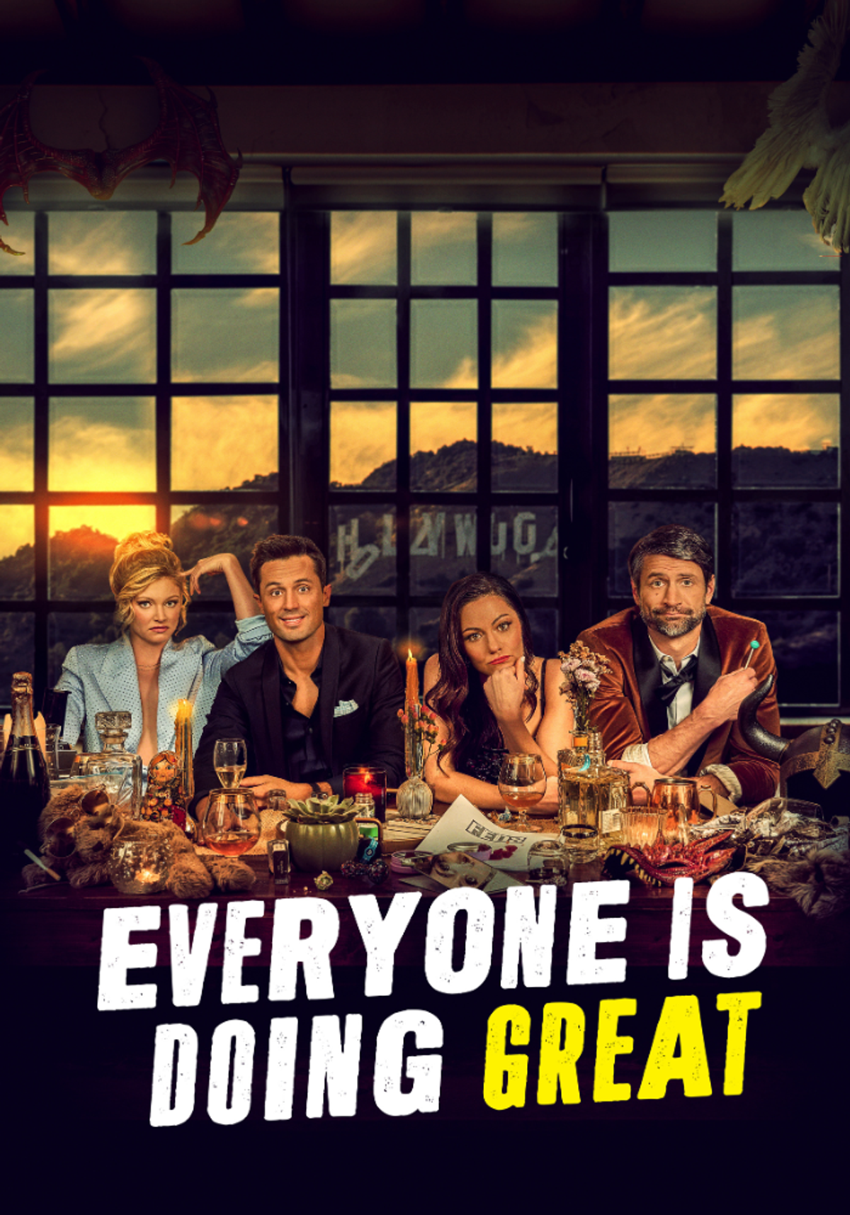 Cast of Everyone Is Doing Great seated around a cluttered dinner table with drinks, candles and props, posed in front of a large window at sunset, with the show’s title displayed across the bottom.