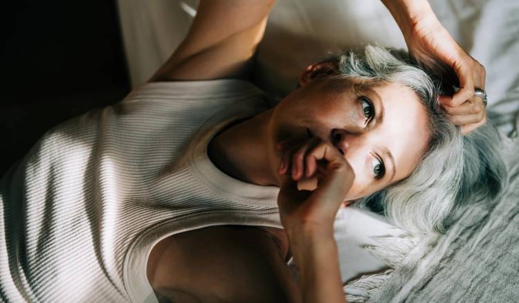 Woman with gray hair lying in bed, looking at the camera with her hand near her mouth