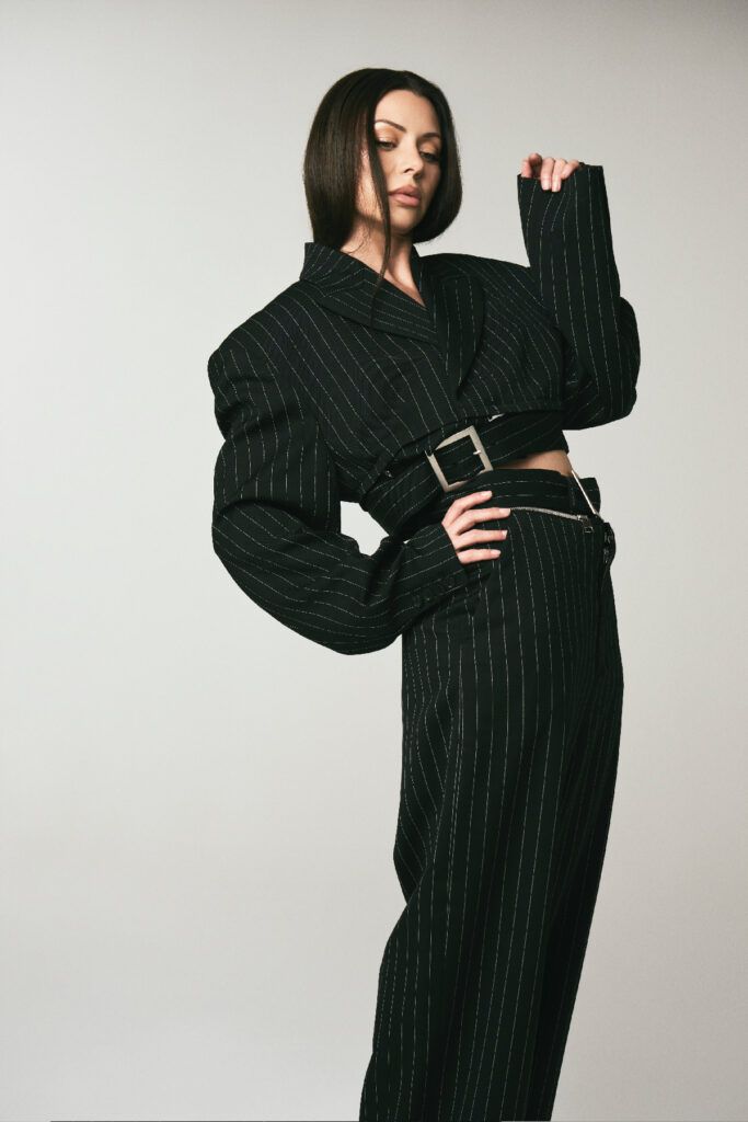 Neraida Bega posing in a black pinstripe cropped blazer and matching trousers, styled with a cinched belt and loose waves