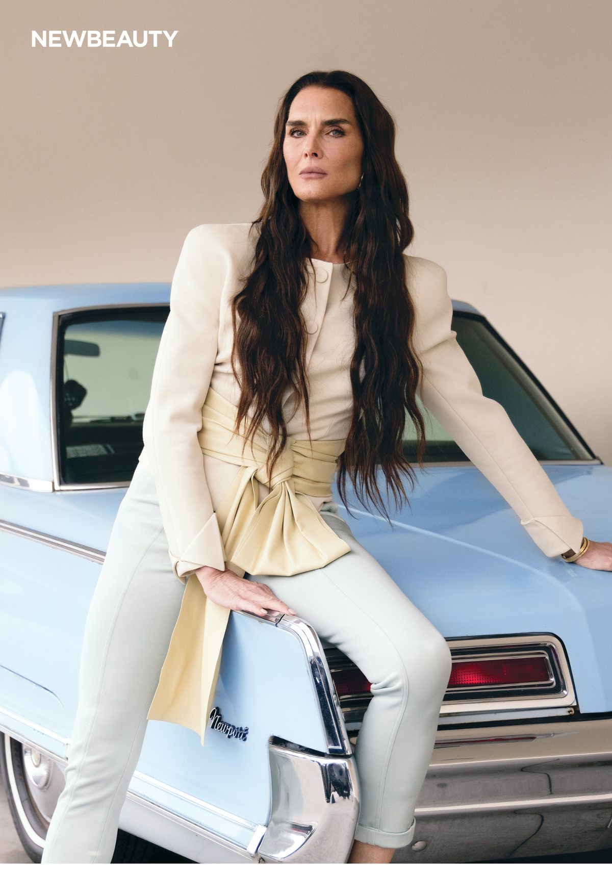 Brooke Shields sits on the trunk of a vintage baby blue Newport car wearing a white blazer, cream top with a yellow wrap detail and white jeans for NewBeauty's summer 2026 cover shoot