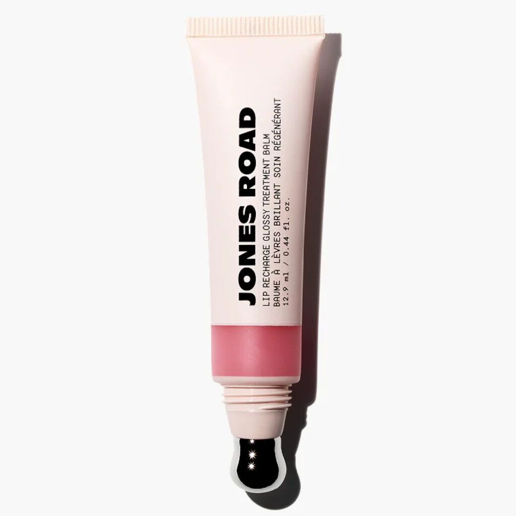 Jones Road Lip Recharge Glossy Treatment Balm in a beige tube with a soft pink-tinted formula visible through a translucent window and a metal applicator tip against a light gray background.