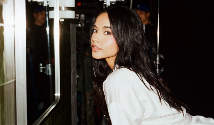 Becky G looks over her shoulder while wearing a white robe backstage, with long dark hair and soft, glowing makeup