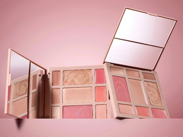 Laura Geller baked face palettes featuring blush, highlighter and eyeshadow in soft pink and bronze tones, displayed against a blush-toned background with open mirrors.