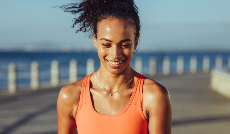 A woman in an orange tank top smiles while sweating after a workout, standing on a seaside boardwalk with the ocean and a railing visible in the background.
