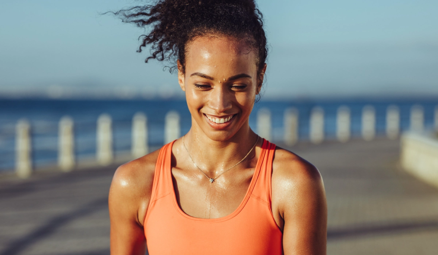 A woman in an orange tank top smiles while sweating after a workout, standing on a seaside boardwalk with the ocean and a railing visible in the background.