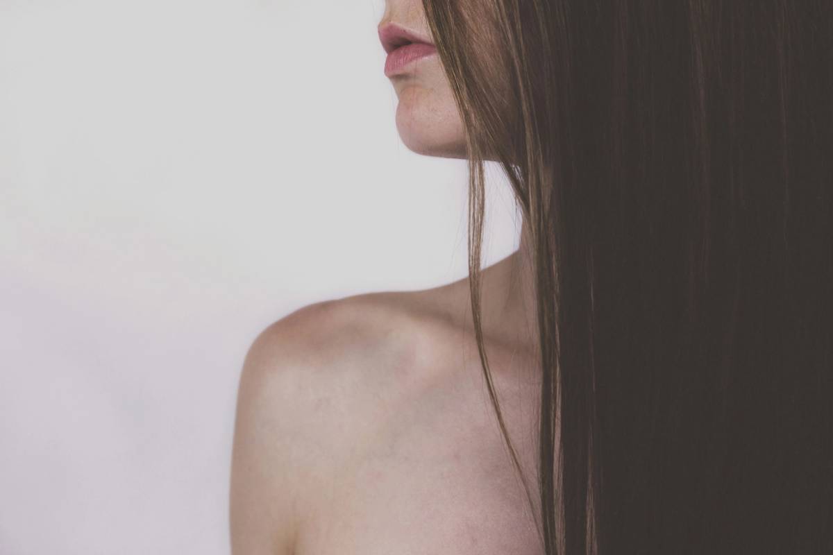 Close-up profile of a woman’s lips, chin and bare shoulder, with long straight brown hair falling past her face against a plain pale gray background.