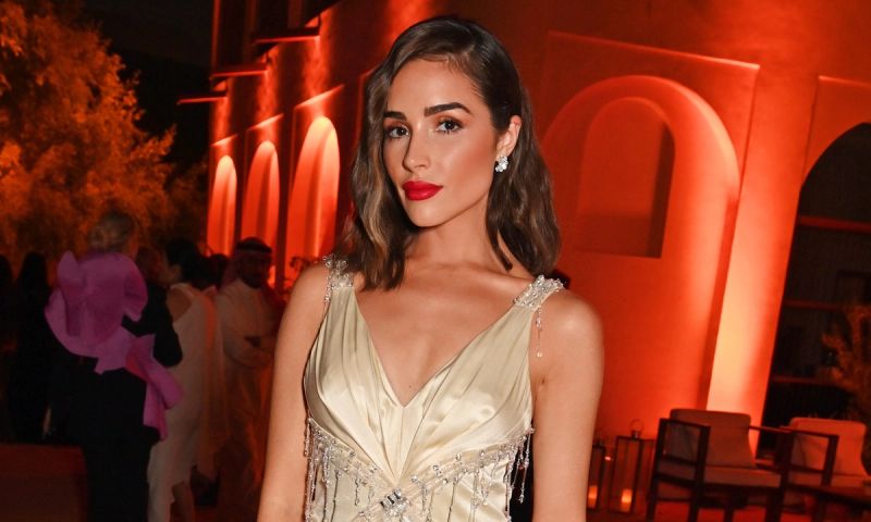 Olivia Culpo wears a gold dress and red lipstick in a red-lit ballroom