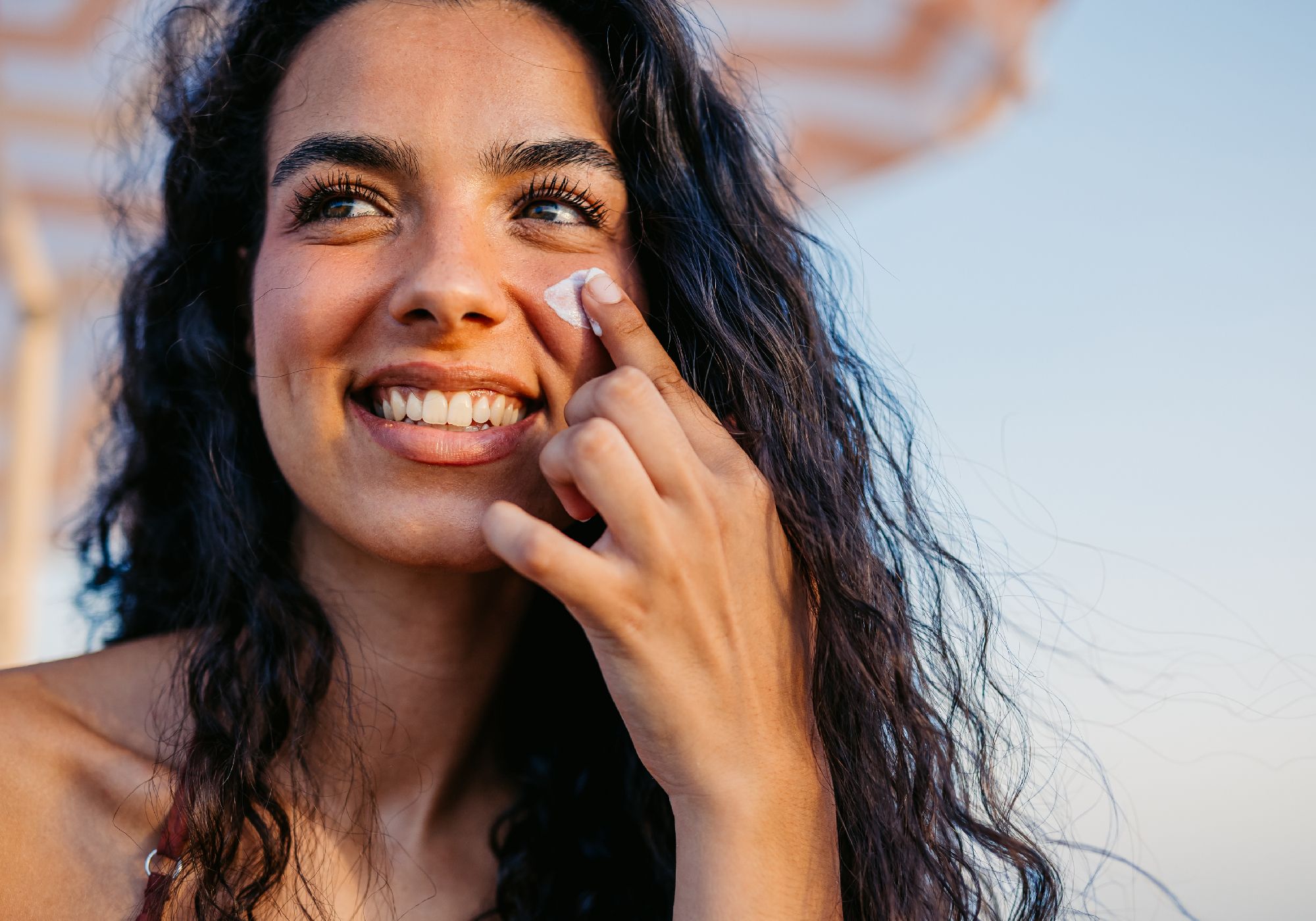 A smiling woman with long, dark curly hair applies a dab of white sunscreen to her cheek with her fingertip, outdoors under a striped umbrella at golden hour
