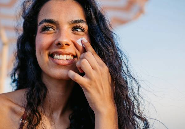 A smiling woman with long, dark curly hair applies a dab of white sunscreen to her cheek with her fingertip, outdoors under a striped umbrella at golden hour