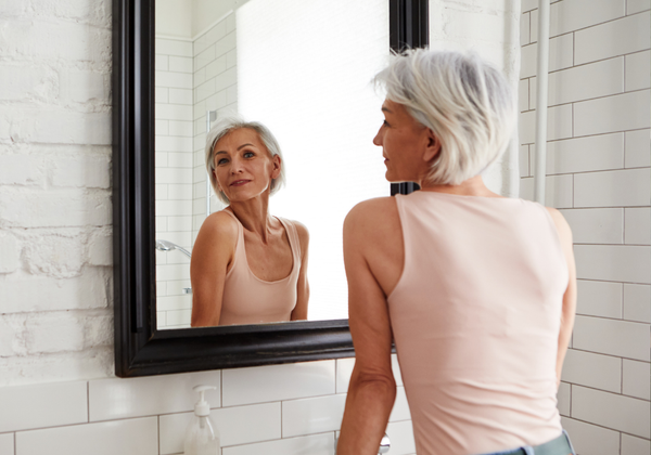 Woman with short gray hair looking in a bathroom mirror, featured in a story about SPF and skin care for mature skin.