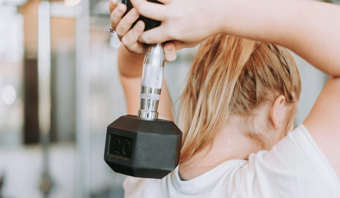 Woman with blonde hair in a white T-shirt performing an overhead triceps extension with a 20-pound dumbbell in a gym.