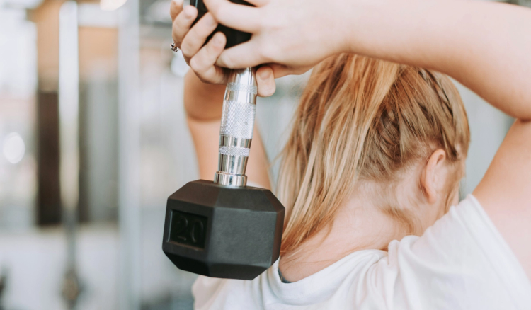 Woman with blonde hair in a white T-shirt performing an overhead triceps extension with a 20-pound dumbbell in a gym.