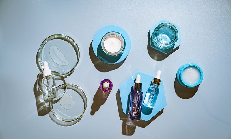 Different glass bottles with serum or oil and cosmetic skin care cream on blue background. Trending shadow. Cosmetic mockup. Top view with copy space. Flat lay.