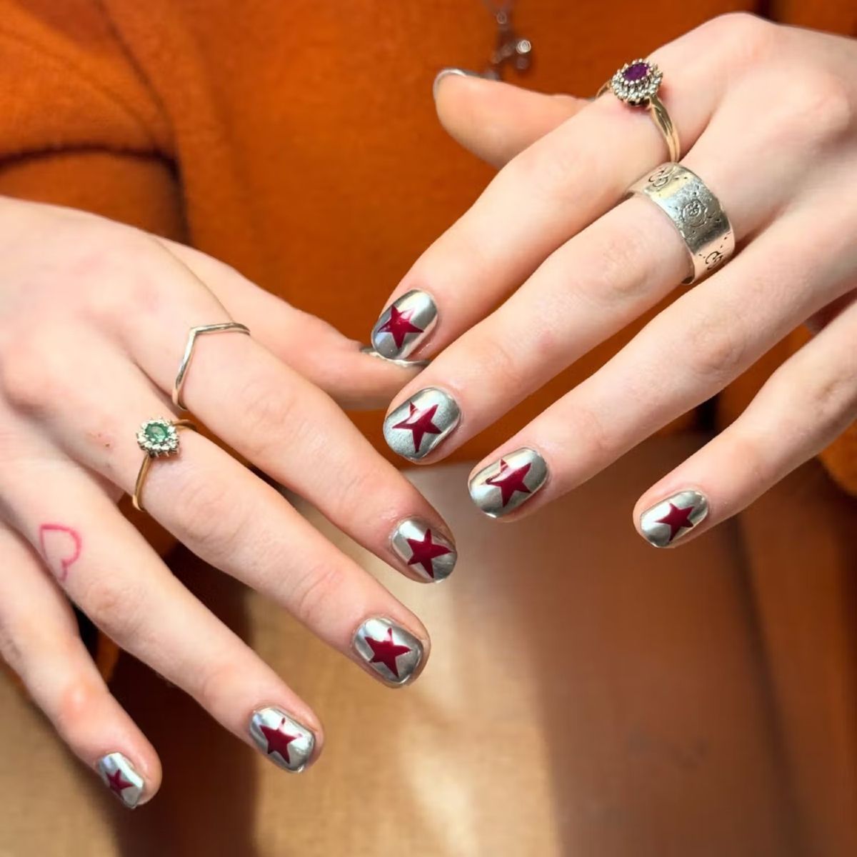 Close-up of hands with short nails painted in a chrome silver finish, each featuring a bold red star design, styled with delicate rings—reminiscent of a Diet Coke–inspired manicure.