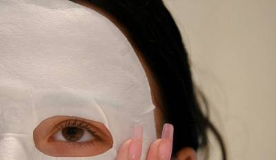 Woman wearing a white sheet mask and touching her face with long pink nails.
