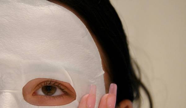 Woman wearing a white sheet mask and touching her face with long pink nails.